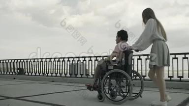 年轻的残疾人<strong>正在</strong>使用轮椅行走，女人<strong>正在</strong>滚动它。 他们一起在河边的公园<strong>里</strong>休息